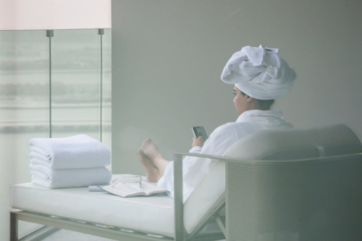 Woman in a white robe with her hair wrapped in a Retreat towel, folded bath towels beside her
