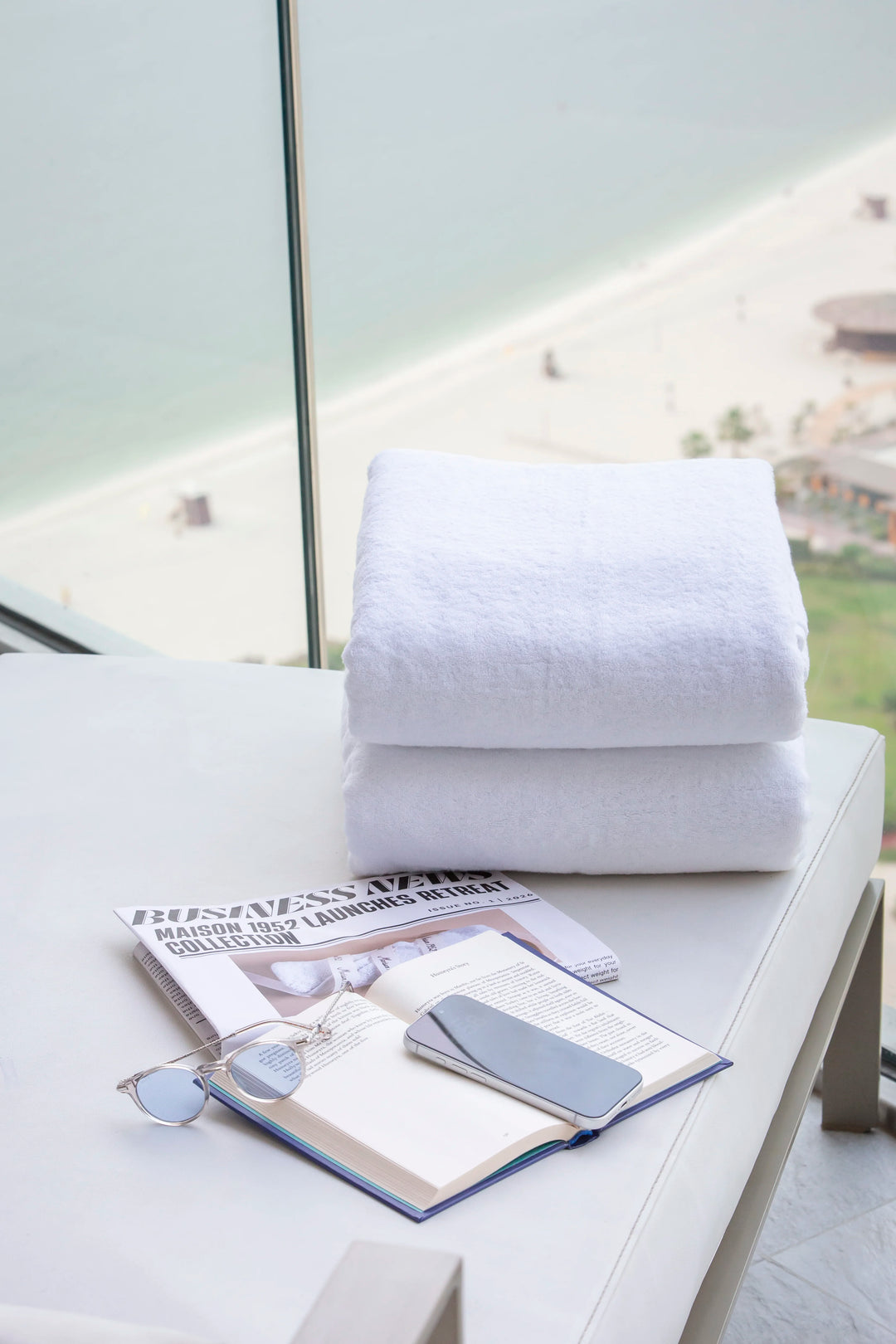 Two folded Retreat white cotton bath towels on a lounger by a window overlooking the beach