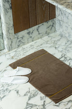 Brown Chateau Turkish cotton bath mat, on a marble bathroom floor with white slippers 