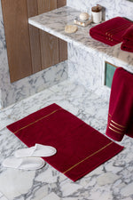 Maroon Chateau Turkish cotton bath mat, on a marble bathroom floor with white slippers 