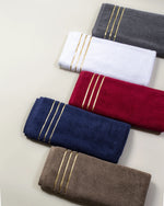 Cinque asciugamani Chateau piegati in cotone turco, colori grigio, bianco, bordeaux, blu navy e marrone, disposti uno accanto all'altro