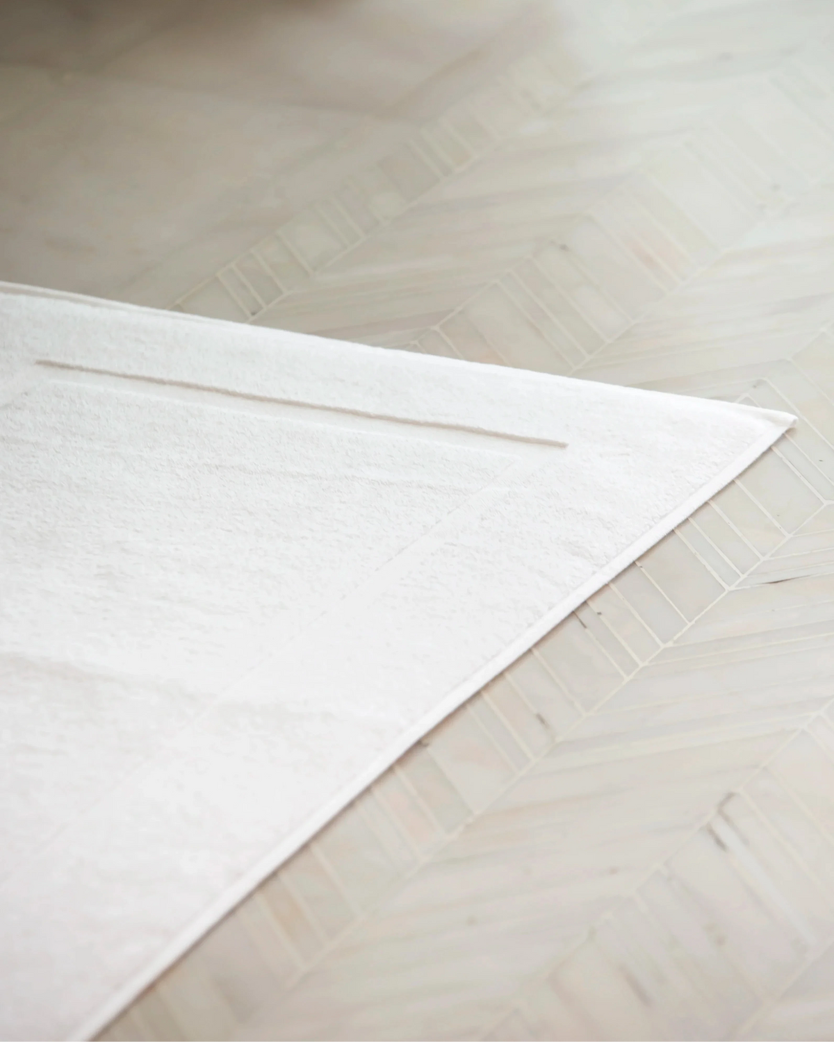 Corner detail of Retreat white cotton bath mat laid flat on a bathroom floor