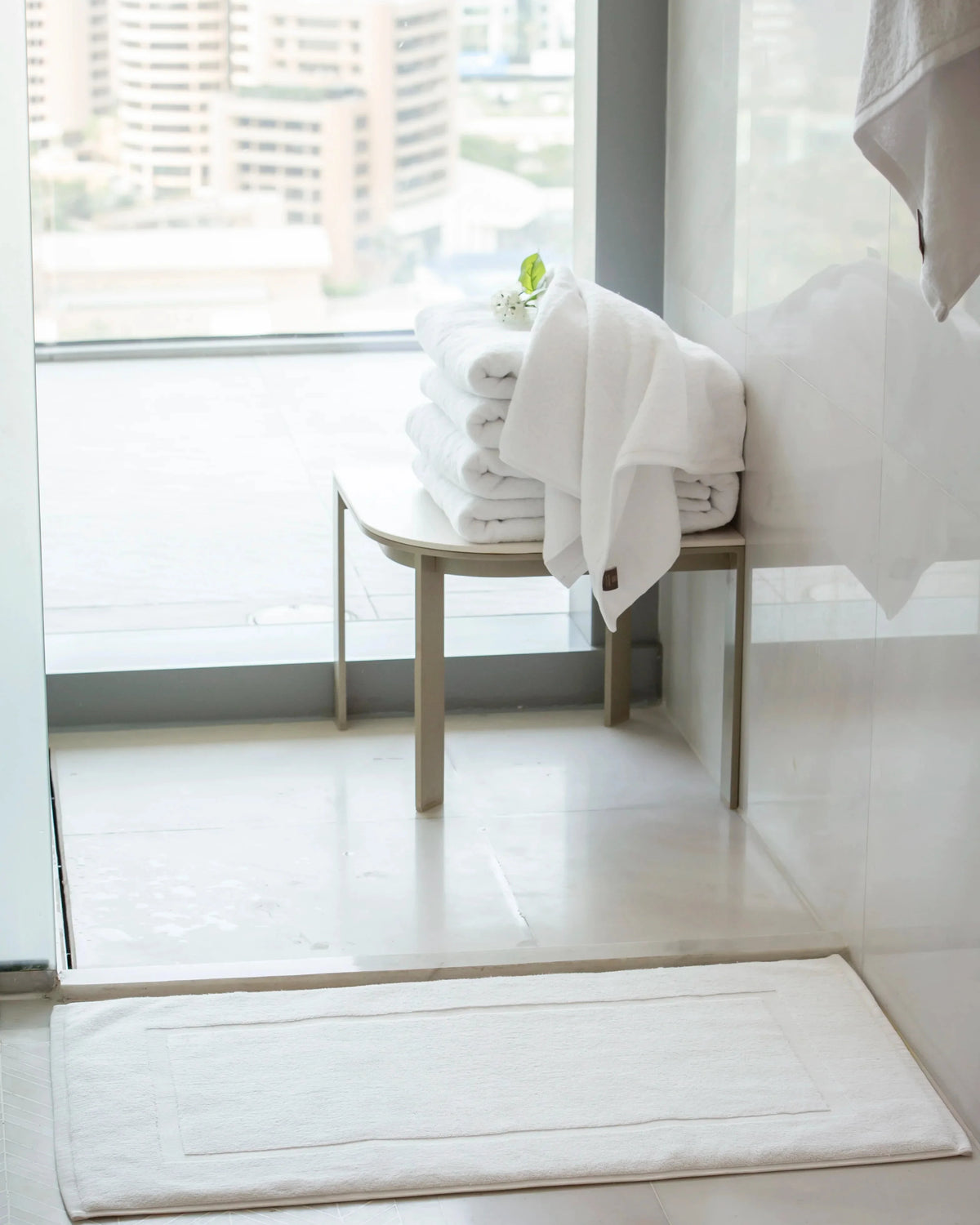 Retreat white cotton bath mat on a bathroom floor with folded white towels by the window